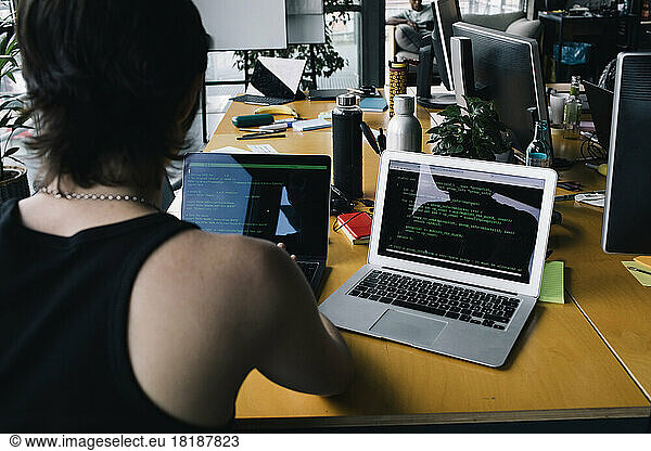 Rear view of non-binary business person coding on laptop at desk in tech-startup company