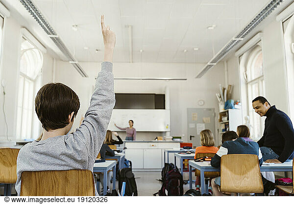 Rear view of schoolboy raising hand while attending lecture in classroom