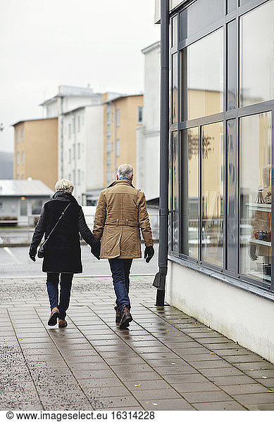 Rear view of senior couple holding hands while walking on sidewalk during winter