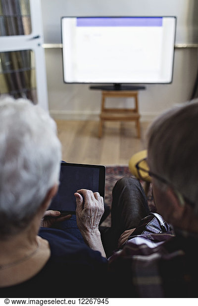 Rear view of senior couple sitting with digital tablet watching TV at home