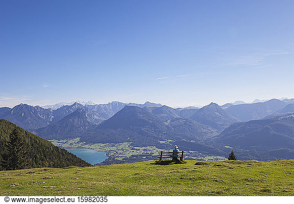 Rear view of senior male hiker sitting on bench while looking at Dachstein Mountains against blue sky