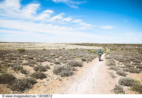 Rear view of senior man riding bicycle on dirt trail amidst semi-arid landscape against sky