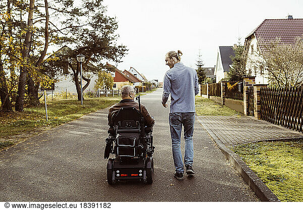 Rear view of senior man with disability in motorized wheelchair by young caregiver walking on road