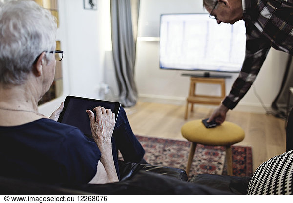 Rear view of senior woman sitting with digital tablet watching TV at home