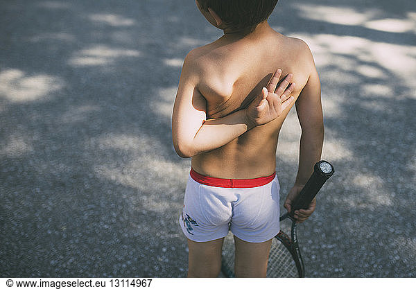 Rear view of shirtless boy holding tennis racket on road