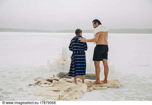Rear view of shirtless man talking with son wearing bathrobe while standing on animal skin at frozen lake