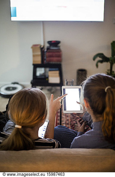 Rear view of sisters using portable information device while sitting in living room at home