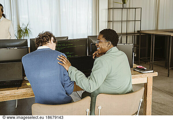 Rear view of smiling female programmer sitting with male colleague at computer desk in office