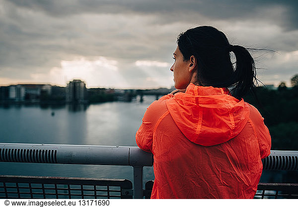 Rear view of sportswoman in raincoat looking at city while standing on footbridge over sea during sunset