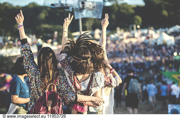 Rear view of three young women standing side by side at a summer music festival wearing feather headdress  arms raised.