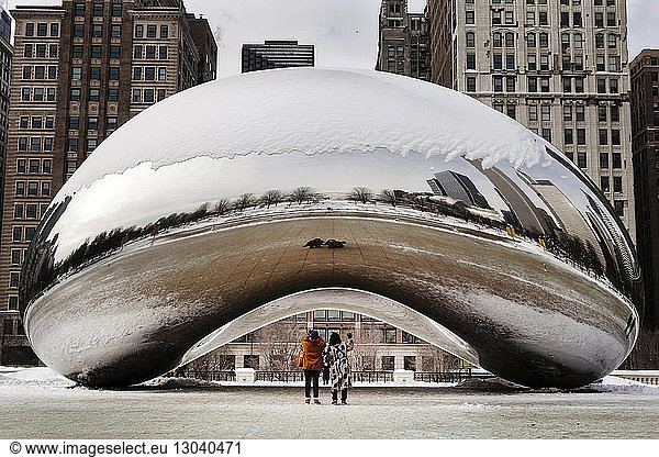 Rear view of tourists standing by snow covered Cloud Gate against buildings at Millennium Park