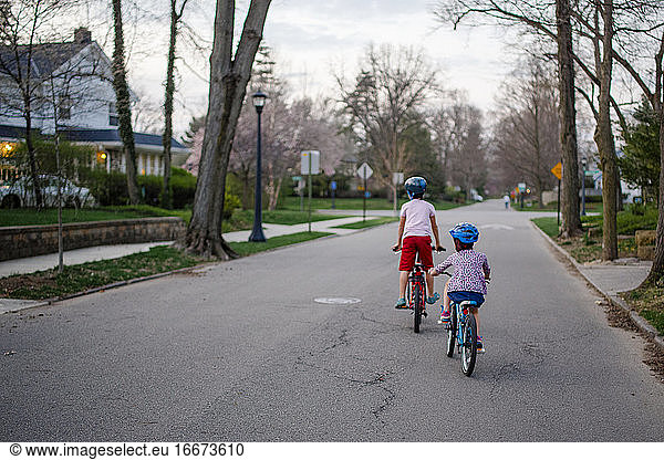 Rear view of two children biking together through neighborhood at dusk