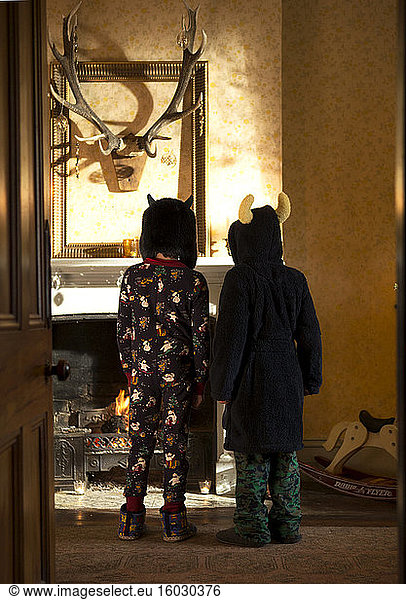 Rear view of two children wearing pyjamas  towelling robe and hats standing in front of a fireplace  hunting trophy on wall.