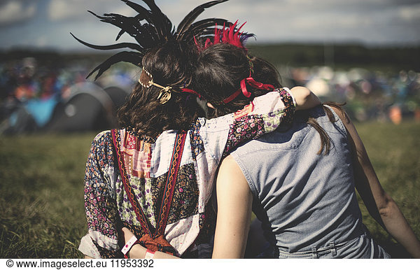 Rear view of two young women at a summer music festival wearing feather headdresses  arm around shoulder.