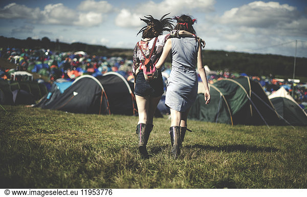 Rear view of two young women at a summer music festival wearing feather headdresses  walking arm in arm towards tents.