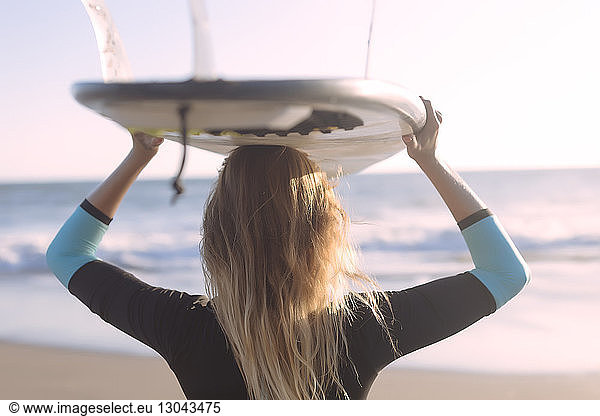 Rear view of woman carrying surfboard on head while standing at beach, Rear view of woman carrying surfboard on head while standing at beach