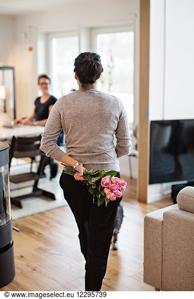 Rear view of woman hiding bouquet while walking towards girlfriend at home