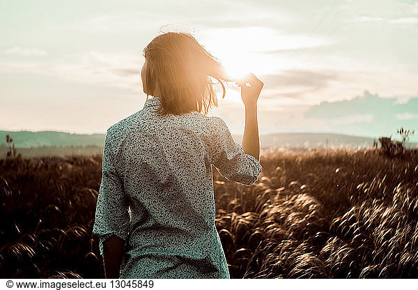Rear view of woman holding hair while standing on field against sky during sunset