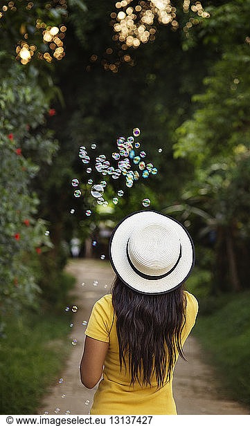 Rear view of woman in sun hat blowing bubbles on footpath