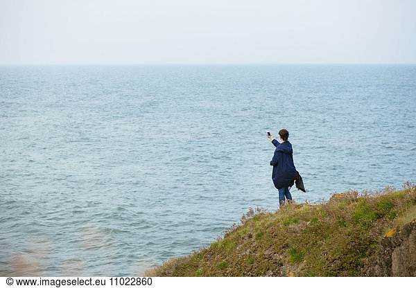 Rear view of woman photographing through smart phone while standing on hill by sea