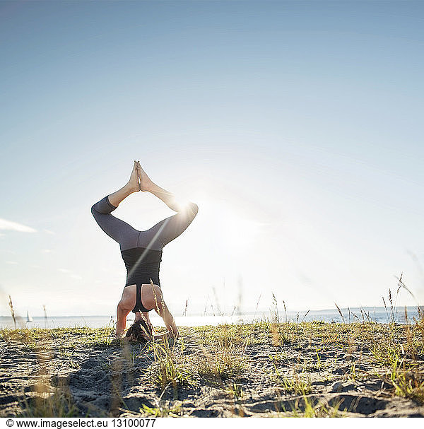 Rear view of woman practicing bound angle headstand pose on sand by sea