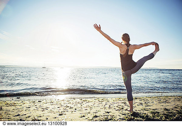Rear view of woman practicing hand to big toe yoga pose at beach during sunny day