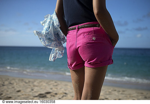 Rear view of woman wearing pink hot pants standing on a sandy beach by the ocean.