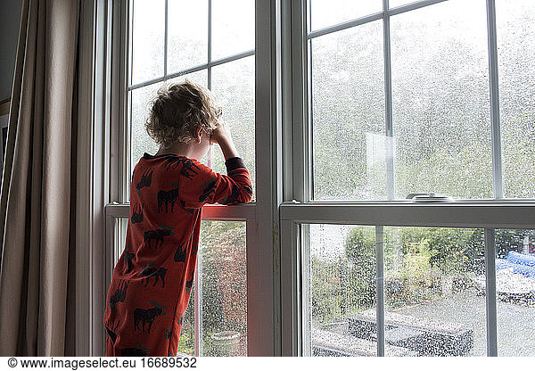 Rear View of Young Boy With Curly Hair Looking Out Rain Covered Window