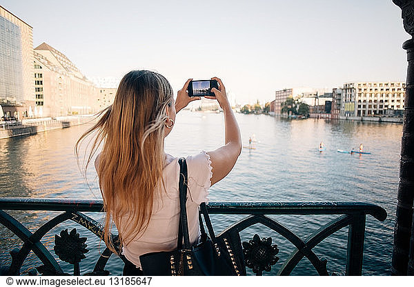 Rear view of young woman photographing river through mobile phone in city