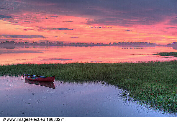 Red canoe floats in Maine tidal marsh amidst dramatic dawn light.