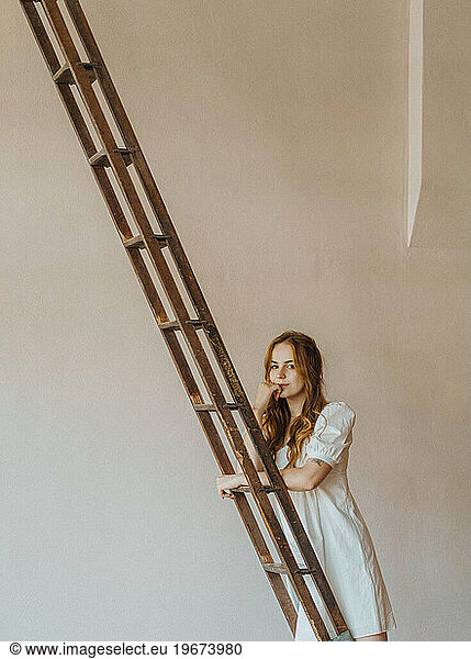 Red-haired model in white poplin dress leans on a vintage ladder.