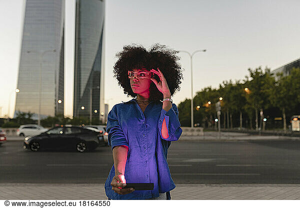 Red light falling on Afro woman face wearing smart glasses holding smart phone on road