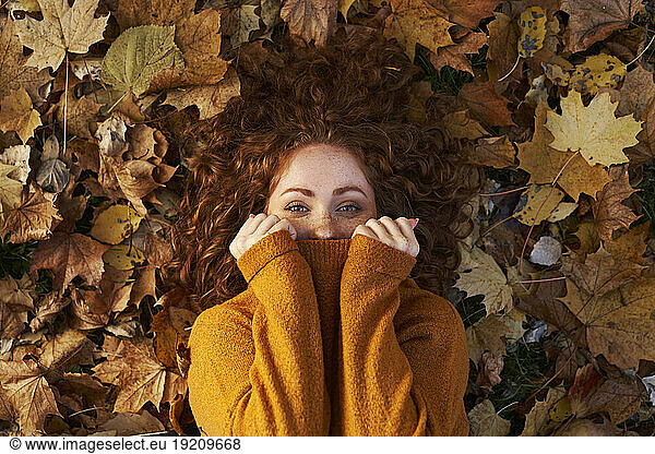 Redhead woman covering face with sweater lying on maple leaves at park
