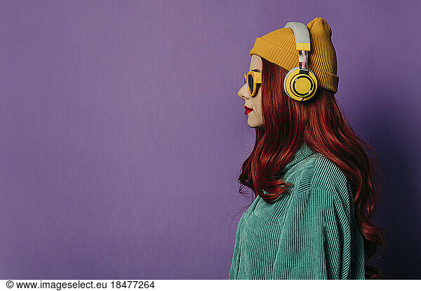Redhead woman listening to music through headphones against purple background