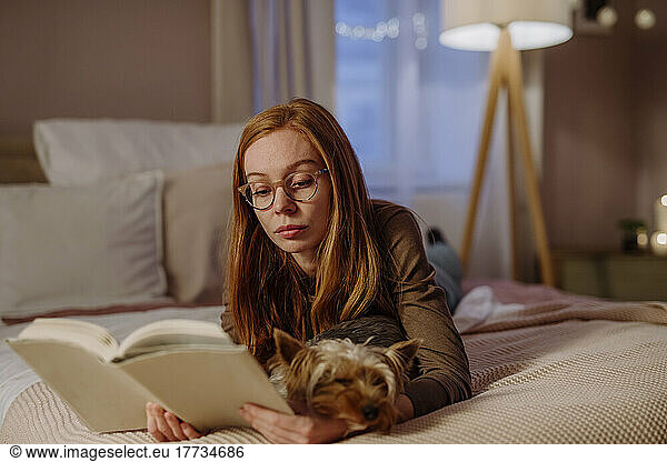 Redhead woman wearing eyeglasses reading book lying with dog on bed at home