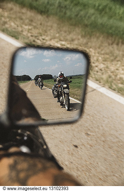 Reflection of friends riding motorcycles on dirt road seen in side-view mirror