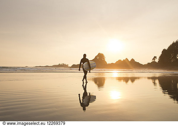 Reflection of man wearing wet suit and carrying surfboard walking along sandy beach at sunset.