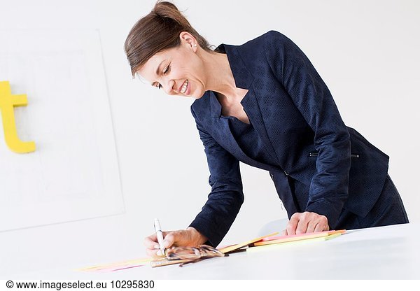 Reife Frau in Geschäftskleidung  die auf Papier schreibt und lächelnd nach unten schaut.