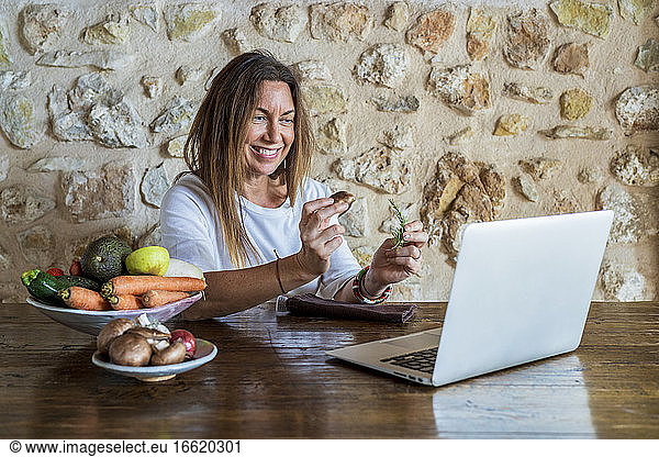 Reife weibliche Food-Influencerin beim Vloggen  während sie Rosmarin und Pilze gegen eine Steinwand hält