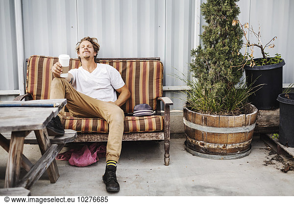 Relaxed carpenter holding disposable cup while sitting on sofa outside workshop