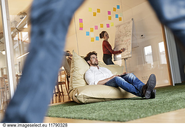 Relaxed man in office sitting in bean bag with meeting in background