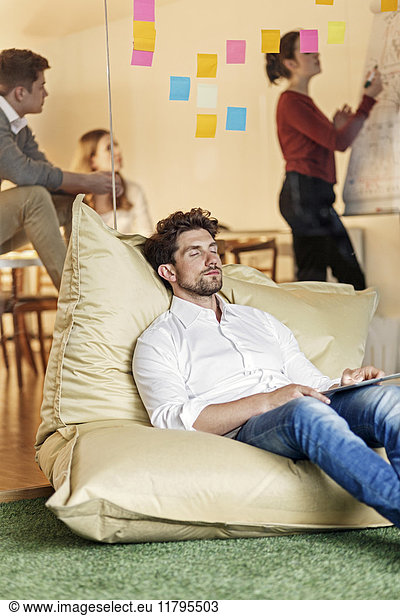 Relaxed man in office sitting in bean bag with meeting in background