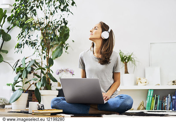Relaxed young woman at home sitting on the floor using laptop and listening to music