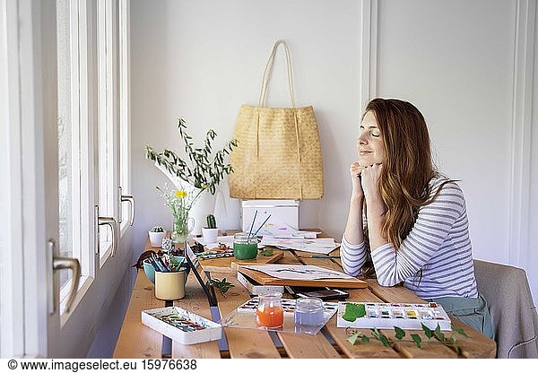 Relaxed young woman sitting with eyes closed while painting on table at home