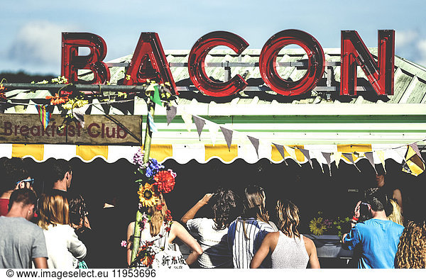 Revellers at a ood stall at a summer music festival red neon sign advertising bacon.