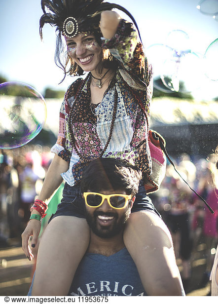 Revellers at a summer music festival young man wearing yellow sunglasses carrying woman wearing feather headdress on his shoulders.