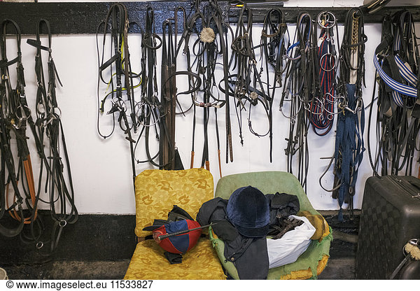 Riding tack on hooks and riding gear on an old armchair in a stable.
