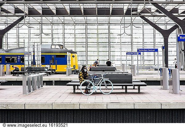 Rotterdam  Niederlande. Mädchen mit blauem Fahrrad wartet auf dem Bahnsteig des Rotterdamer Centraal-Bahnhofs auf ihren Anschlusszug  während sie wegen der Corona-Krise ihren vorgeschriebenen Mundschutz aufsetzt.