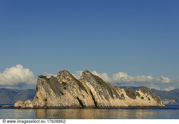 Rugged landforms along the ocean coastline under a blue sky with cloud resting over mountaintops  Komodo National Park; East Nusa Tenggara  Indonesia