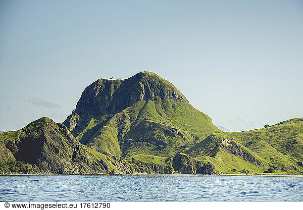 Rugged landforms covered in green foliage along the ocean coastline under a blue sky  with birds standing on the peaks  Komodo National Park  East Nusa Tenggara  Indonesia